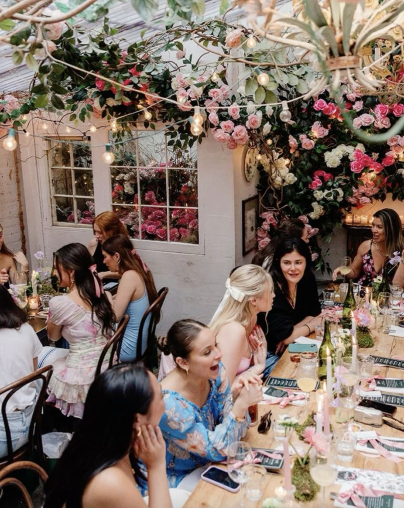 Elegant outdoor dinner party women at long table under pink rose floral arch string lights candles romantic garden gathering aesthetic showcasing Modern PR Isnt Just Press.