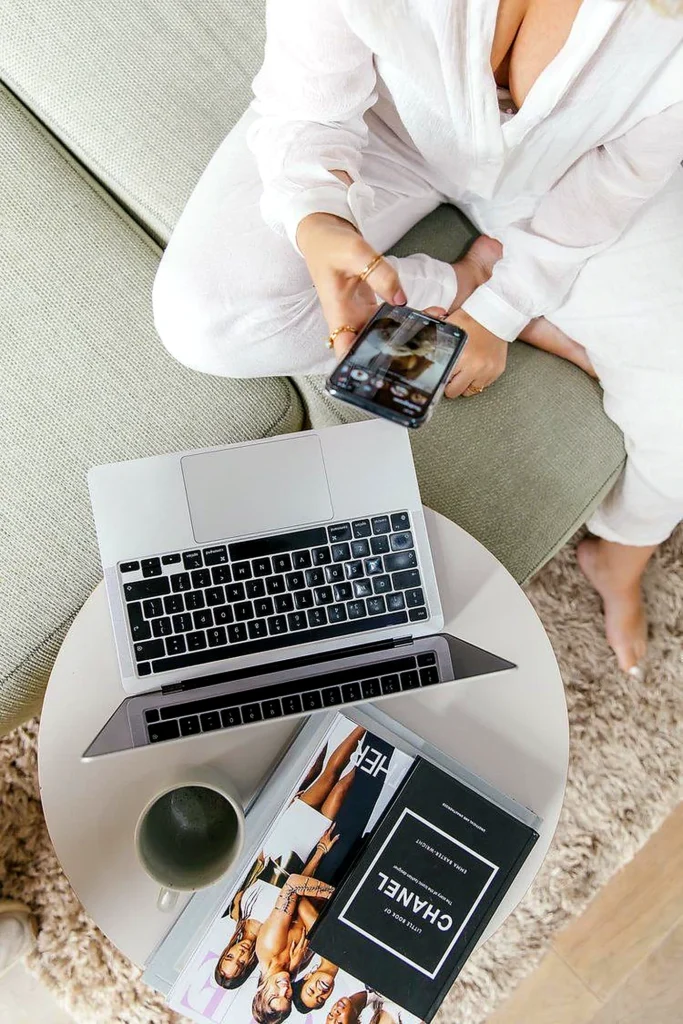 Woman using a phone beside a laptop, coffee, and fashion magazines Suggesting How Consumers Discover Beauty Brands