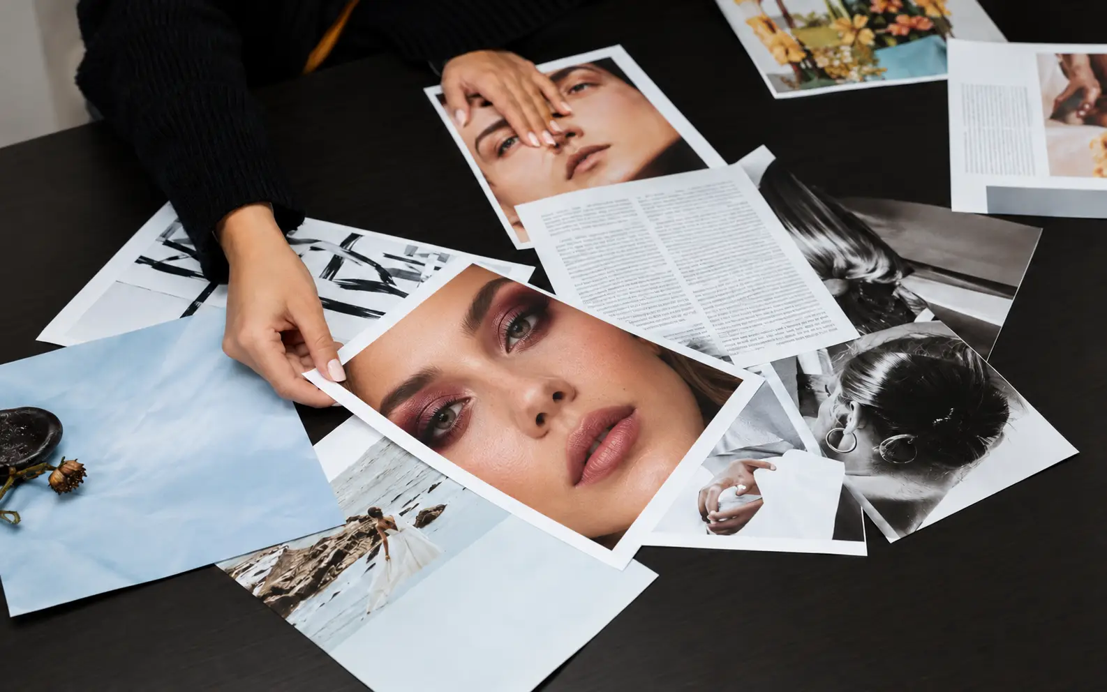 Editorial Roundups Hands arranging beauty and fashion magazine pages on a dark wooden desk.