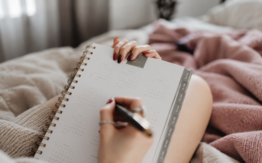 Hands holding a spiral-bound planner and writing with a pen beside a pink blanket.