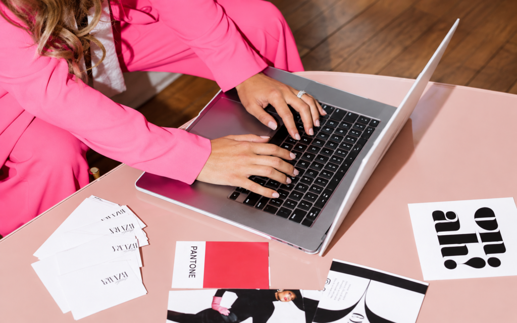 Person in a bright pink shirt typing on a laptop beside printed design materials.