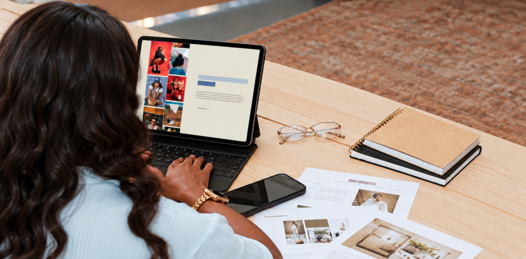 Person using a tablet and keyboard beside printed photo layouts, reading glasses, and a smartphone.
