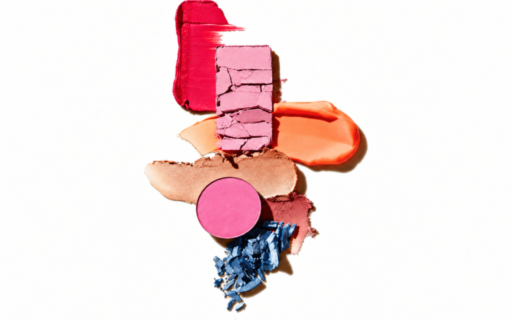 Pink and coral makeup swatches with broken pressed powders and blue pigment on a white background.