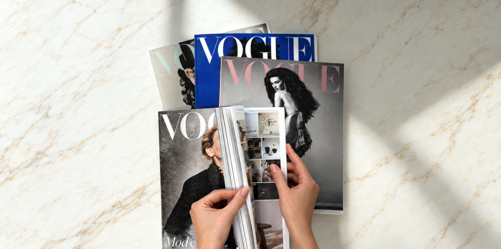 Hands browsing an open fashion magazine in front of a stack of Vogue issues.