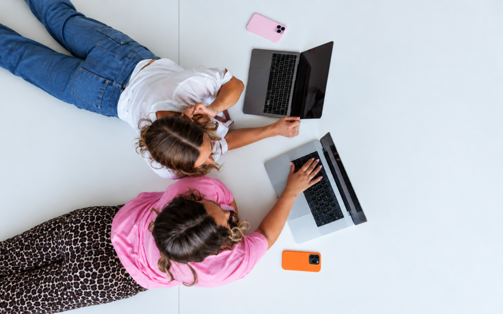 Overhead view of two people in white and pink tops working on separate laptops at a white desk.