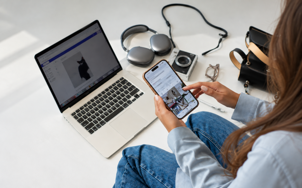 Person in a blue shirt holding a smartphone beside a silver laptop and over-ear headphones.